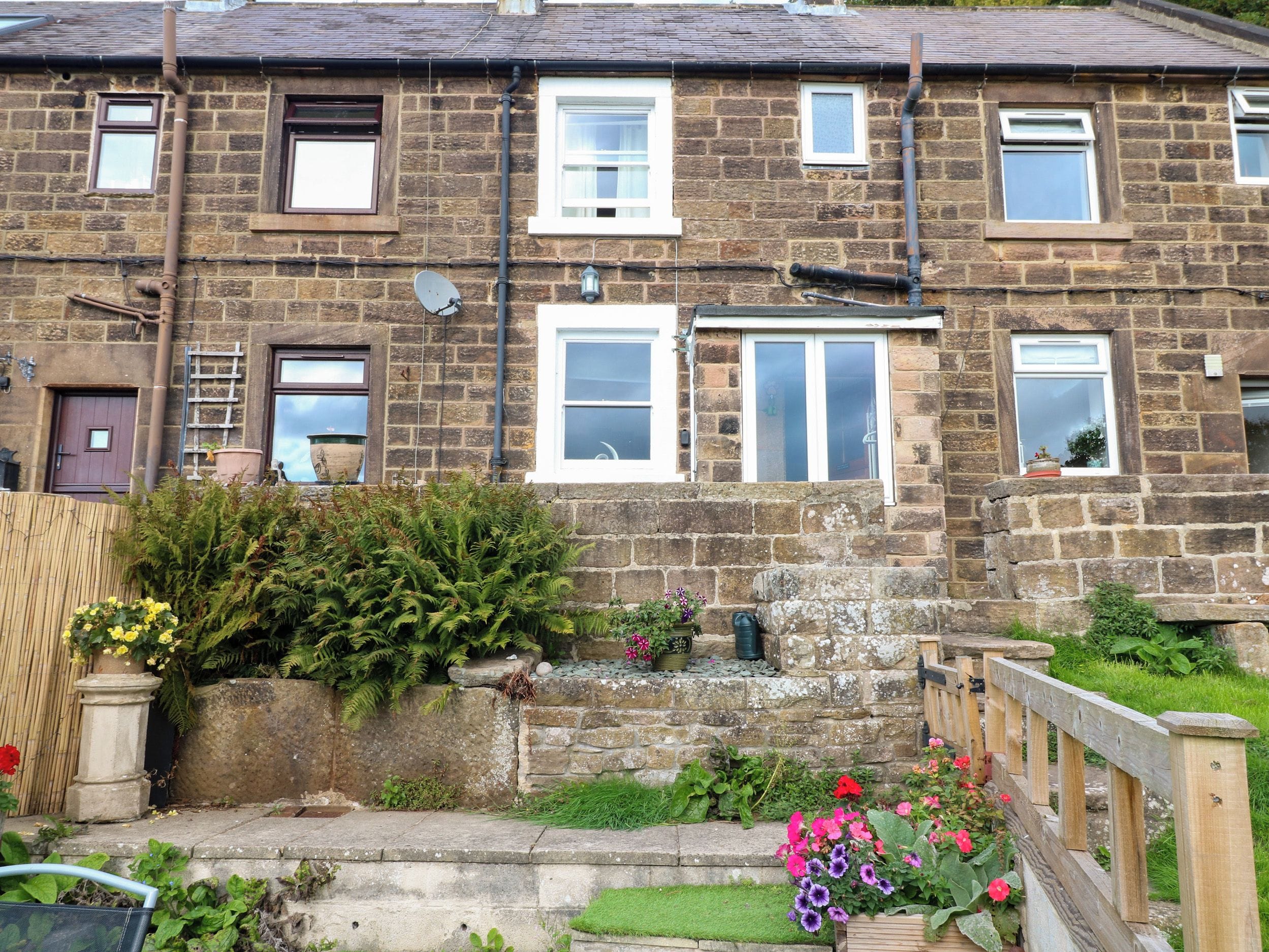 A two-story stone house with multiple windows, a satellite dish, and a small garden with various plants and flowers.