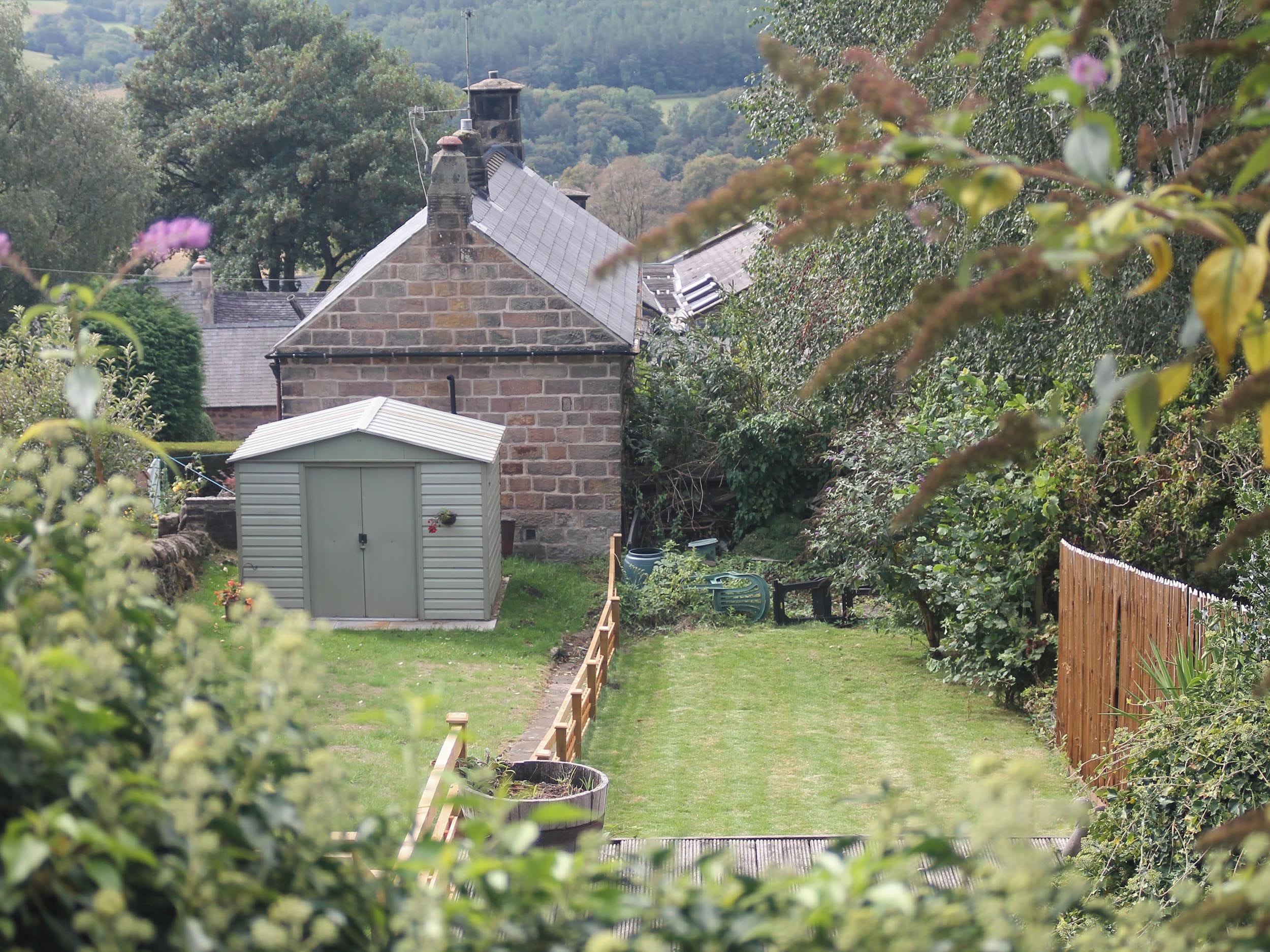 A stone house with a small shed, surrounded by a wooden fence and lush greenery.