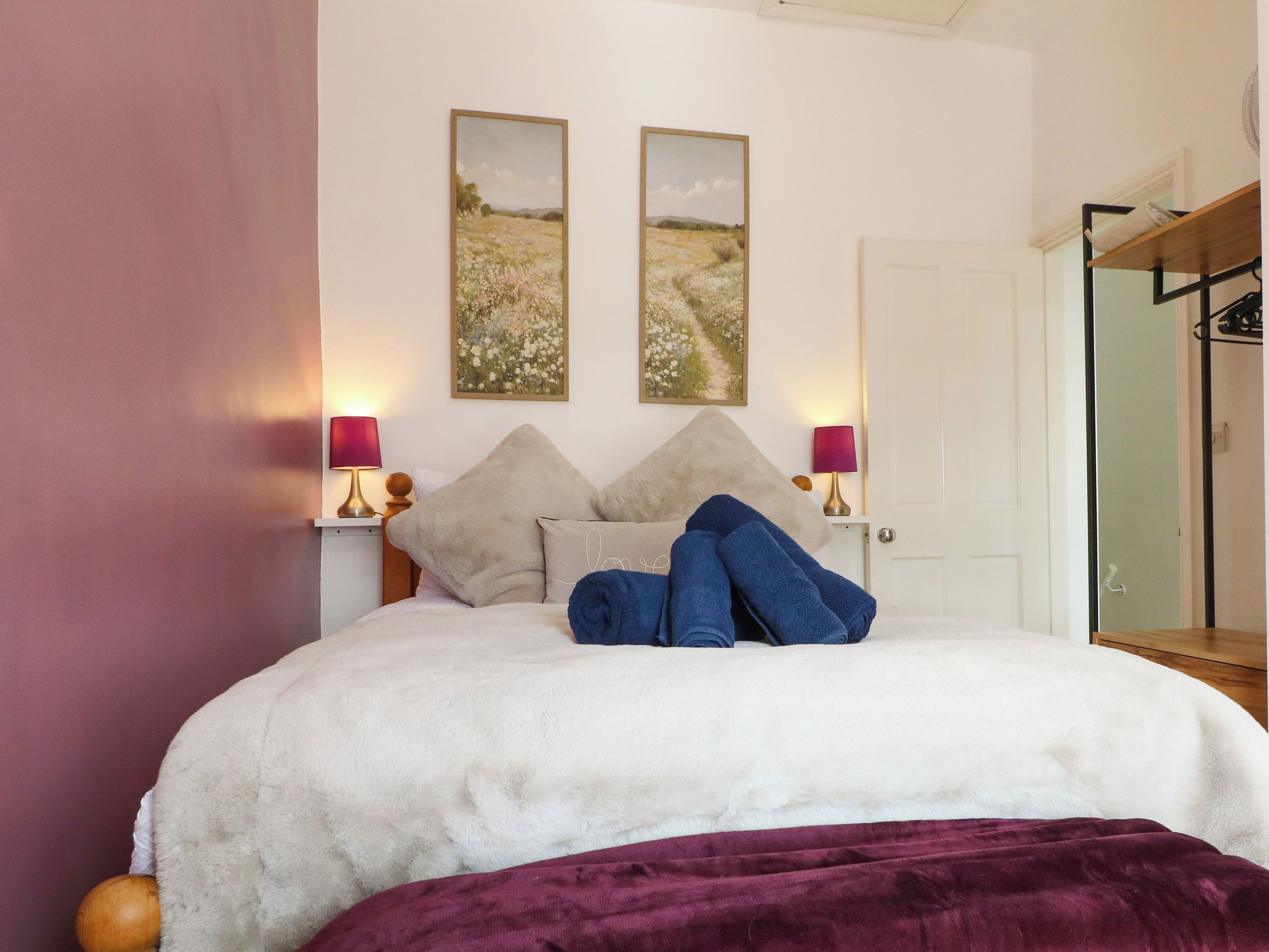 A bed with a white duvet, two pillows, and a purple blanket. There are two wall-mounted lamps with pink shades.