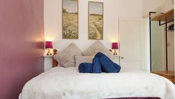 A bed with a white duvet, two pillows, and a purple blanket. There are two wall-mounted lamps with pink shades.
