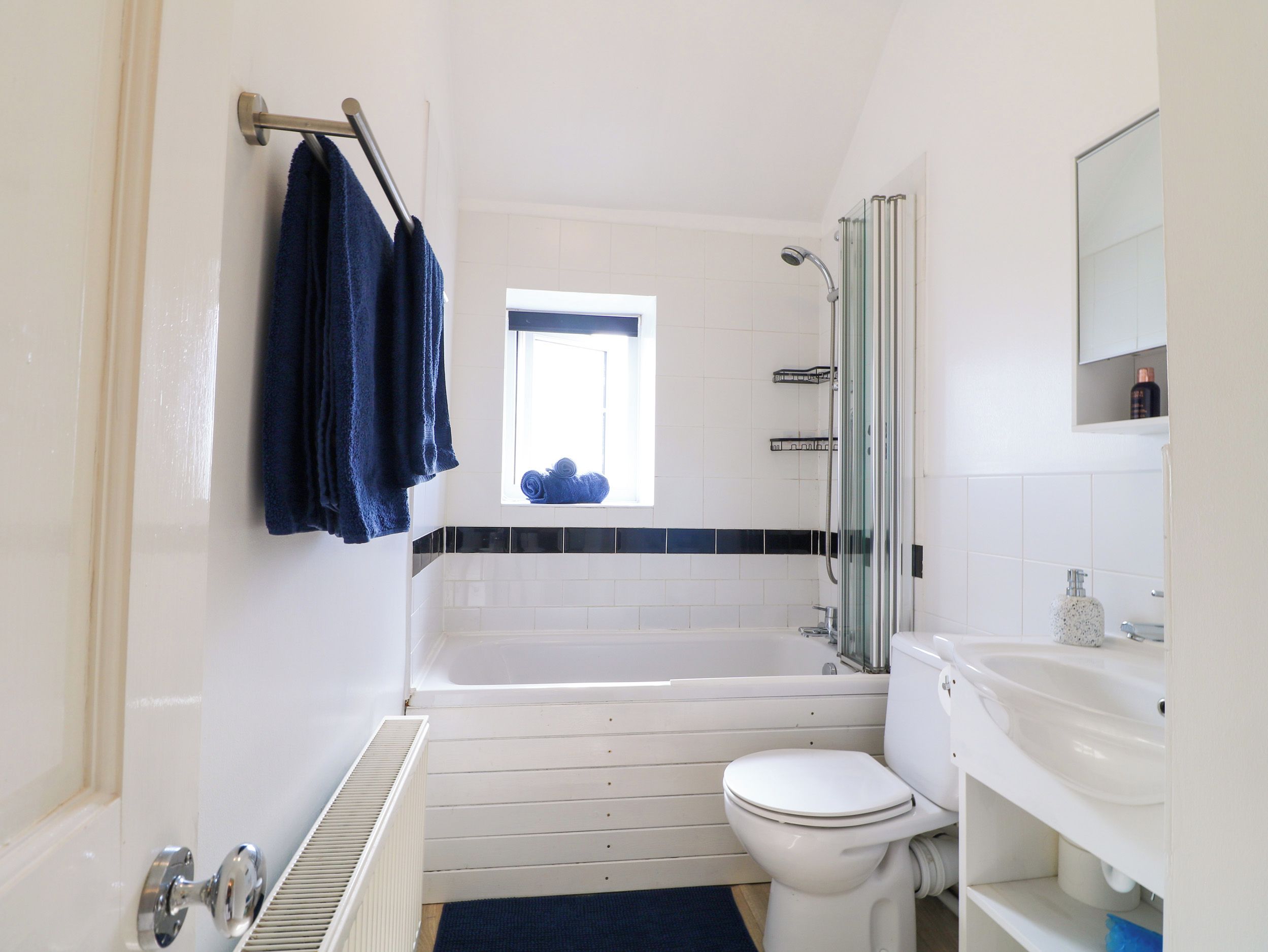 Un baño con una bañera blanca, un inodoro blanco, una cabina de ducha con paredes de cristal y una ventana sobre la bañera.