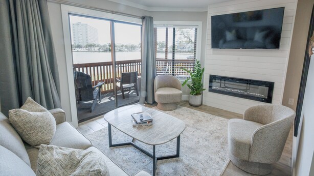 A living room with a sofa, armchair, coffee table, TV, fireplace, and a view of a body of water through large windows. - 112 Arrowhead Unique Lakefront 2br At Kingston 2 Bedroom Condo (Myrtle Beach)