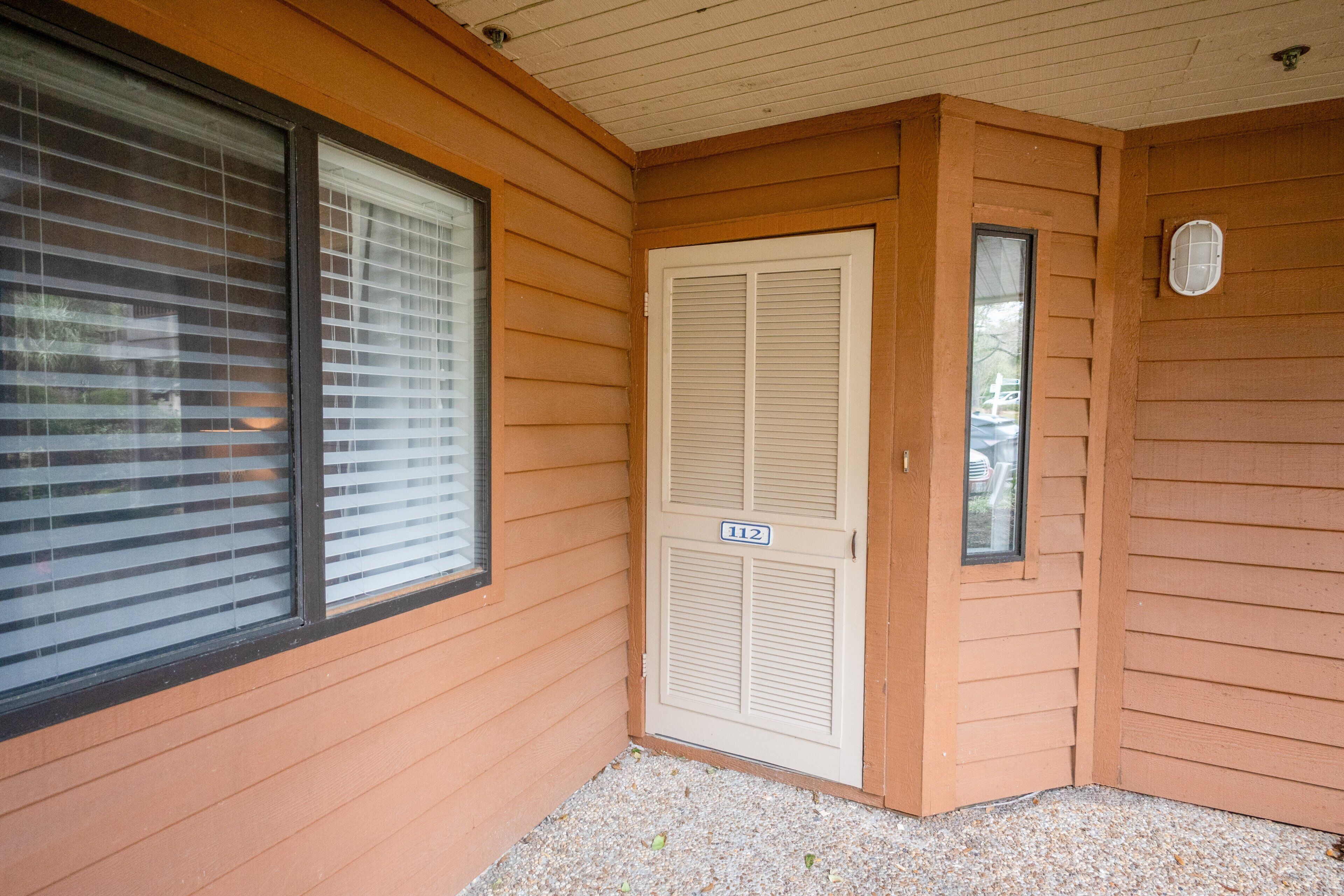 A corner of a building with wooden siding, a closed door with a number plate, and a window with blinds.