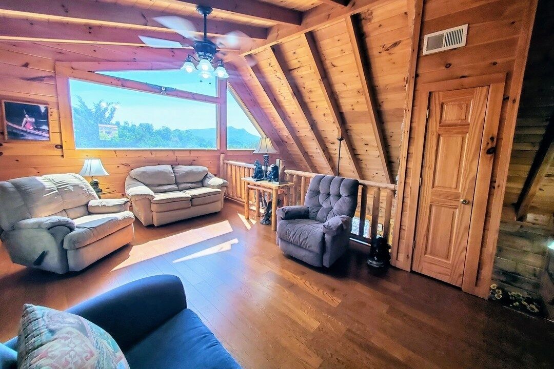 A cozy loft-style room with a wooden ceiling, a large window, and a seating area with a sofa and armchairs.