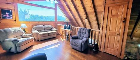 A cozy loft-style room with a wooden ceiling, a large window, and a seating area with a sofa and armchairs.