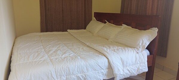 Una cama con colcha y almohadas blancas, cabecera de madera y pared lisa con panel de madera.