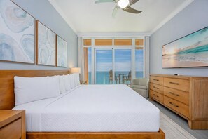 Habitación de hotel con una cama grande, cabecera de madera, ventilador de techo, televisor, cómoda de madera y balcón con vista al océano.
