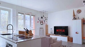 Et moderne køkken med en central ø, et spiseområde med en sofa, et fjernsyn der viser 'NETFLIX' og udsigt til det fri gennem store vinduer.