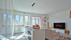 Et moderne køkken med en central ø, et spiseområde med sofa, et fladskærms-tv og et stort vindue.