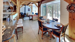 A cozy cabin interior with a dining area, kitchen, and living space, featuring wooden beams, large windows, and a fireplace.