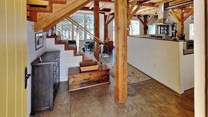 Un escalier en bois menant à un deuxième étage, un coin cuisine avec un comptoir et des appareils électroménagers, et une vue sur l’extérieur grâce à de grandes fenêtres.