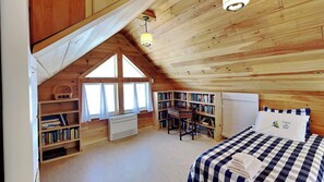 A cozy attic bedroom with a sloped ceiling, wooden bookshelves, a desk, a bed with checkered bedding, and a window with curtains.