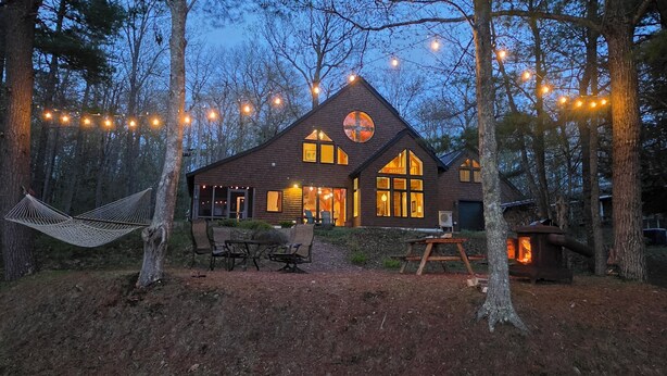 Un chalet rustique doté d’une grande fenêtre circulaire, entouré d’arbres et illuminé de guirlandes lumineuses.