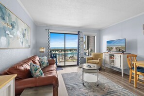 A modern living room with a leather sofa, a round coffee table, a TV, and a view of a beach and pool. - Oceanfront 1 BR Condo | Myrtle Beach Resort A0105 (Myrtle Beach)