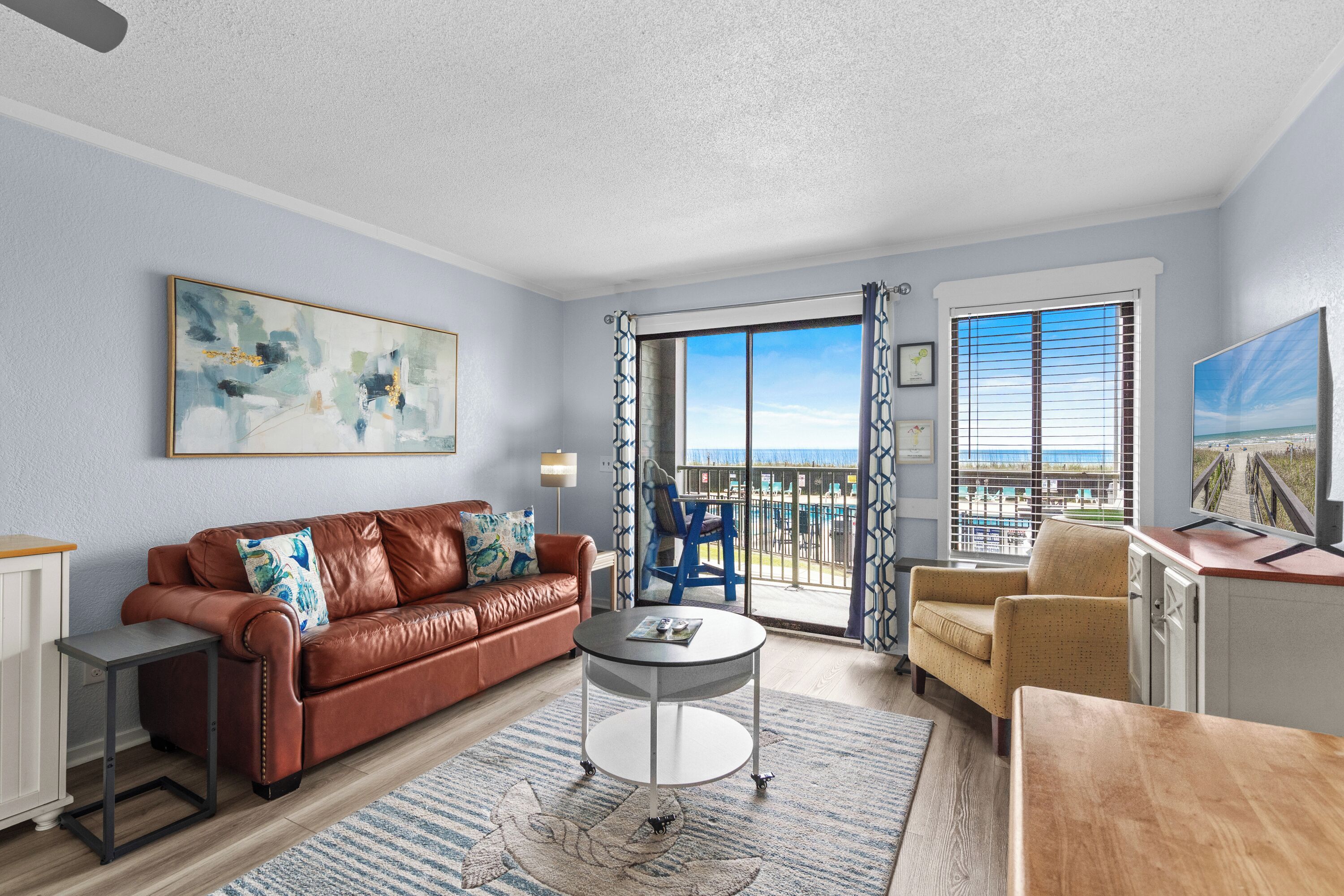 A living room with a brown leather sofa, a small round table, a blue chair, and a view of the beach through a large window.