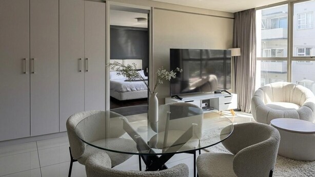 Ein modernes Wohnzimmer mit einem Glastisch, einer Couch, einem Fernseher und einem Bett im Hintergrund.