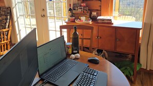 Ein Interieur einer Hütte mit einem Laptop, einer Tastatur und einem Notizblock auf einem Holztisch, einer Flasche und einer Lebensmitteldose auf einem kleinen Tisch sowie einem Blick ins Freie durch ein Fenster.