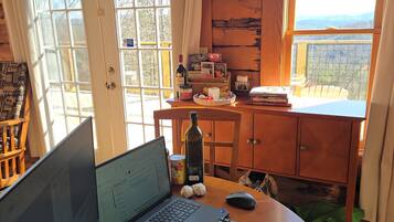 Interior de una cabaña con una computadora portátil, teclado y libreta sobre una mesa de madera, una botella y una lata de comida sobre una mesita, y vista al exterior a través de una ventana.