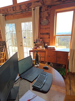 L’intérieur d’une cabane avec un ordinateur portable, un clavier et un bloc-notes sur une table en bois, une bouteille et une conserve sur une petite table, et une vue sur l’extérieur par une fenêtre.