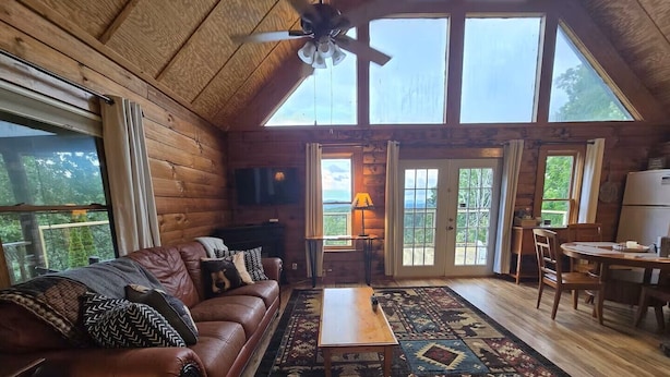 Ein gemütliches Blockhaus-Interieur mit Ledersofa, Esstisch, Fernseher und einem großen Fenster mit Blick auf Grünfläche.