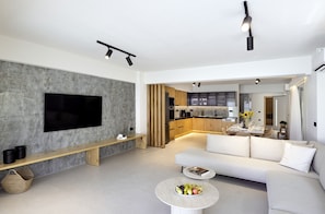 A modern living room with a flat-screen TV, a sofa, a coffee table with fruit, and a kitchen in the background. - Atlantis Homes 1, By Hellocrete (Rethymno)