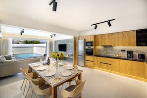 A modern kitchen with wooden cabinets, a dining area with a table set for four, and a large window overlooking a pool. - Atlantis Homes 1, By Hellocrete (Rethymno)