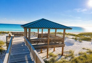 Ein hölzerner Strandpavillon mit Dach, ein Holzdeck davor und ein sandiger Strand mit Gras im Vordergrund.