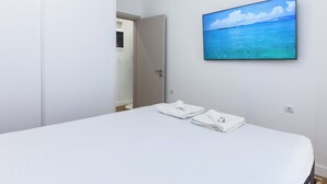 A hotel room with a bed, a wall-mounted TV displaying a beach scene, and a door leading to another room. - Athens Riviera 2BR Apartment by ΜΡS (Kallithea)
