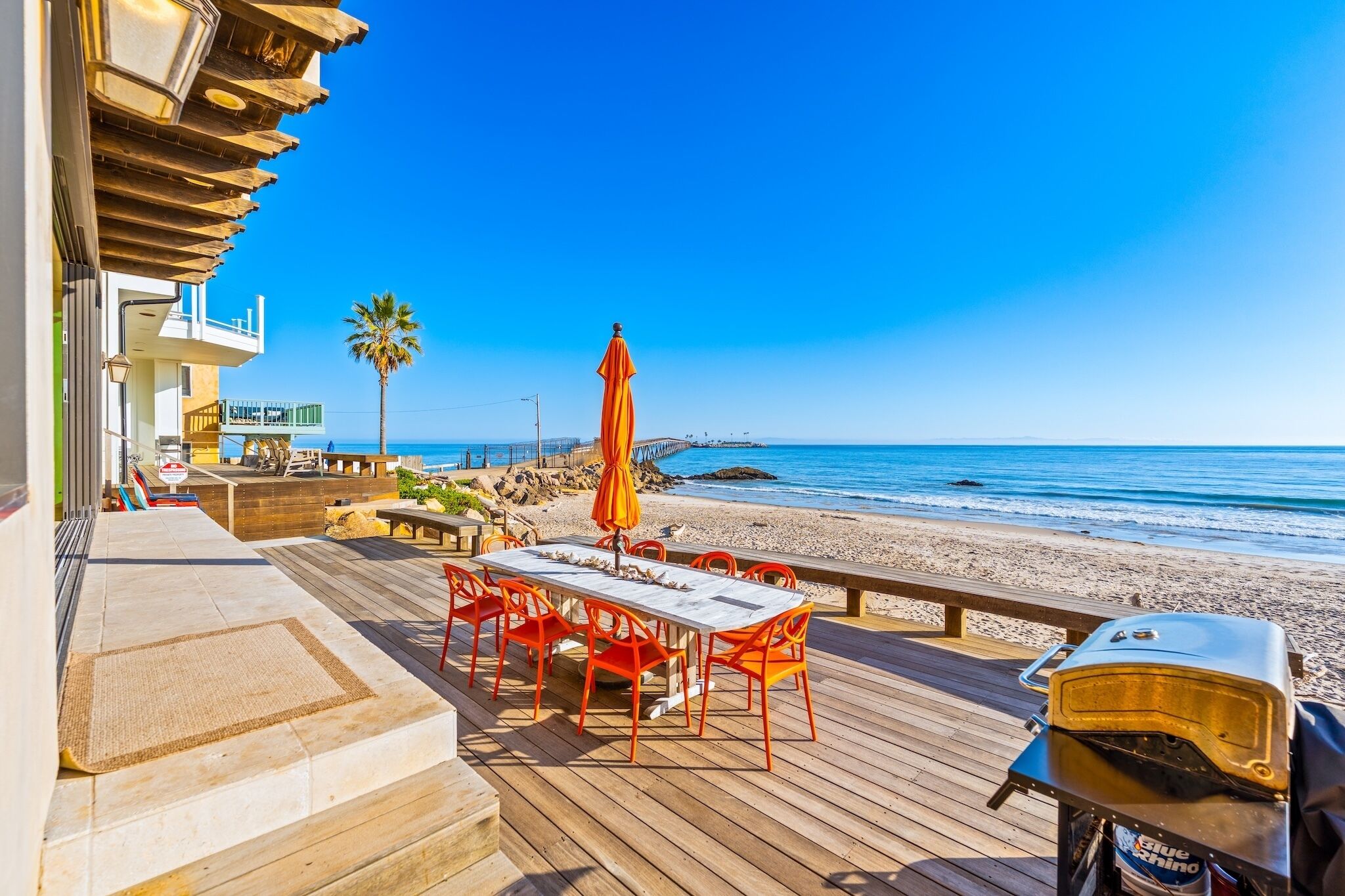 Eine Holzterrasse mit Tisch und Stühlen, einem Grill und Blick auf Strand und Meer.
