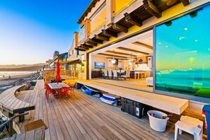 Une terrasse en bois avec des sièges d’extérieur, une vue sur la plage et une grande fenêtre donnant sur l’océan.