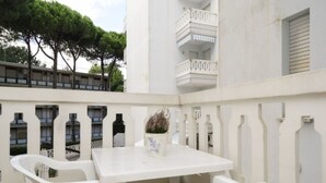 Un espacio exterior con mobiliario blanco, una mesa, sillas, una planta en maceta y un balcón con barandillas.