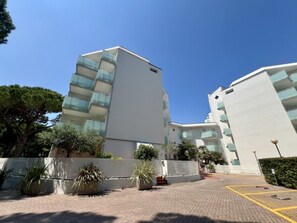 A modern multi-story building with balconies, a paved entrance area, and surrounding greenery. - Cozy Accommodation With Shared Pool (Lignano Sabbiadoro)