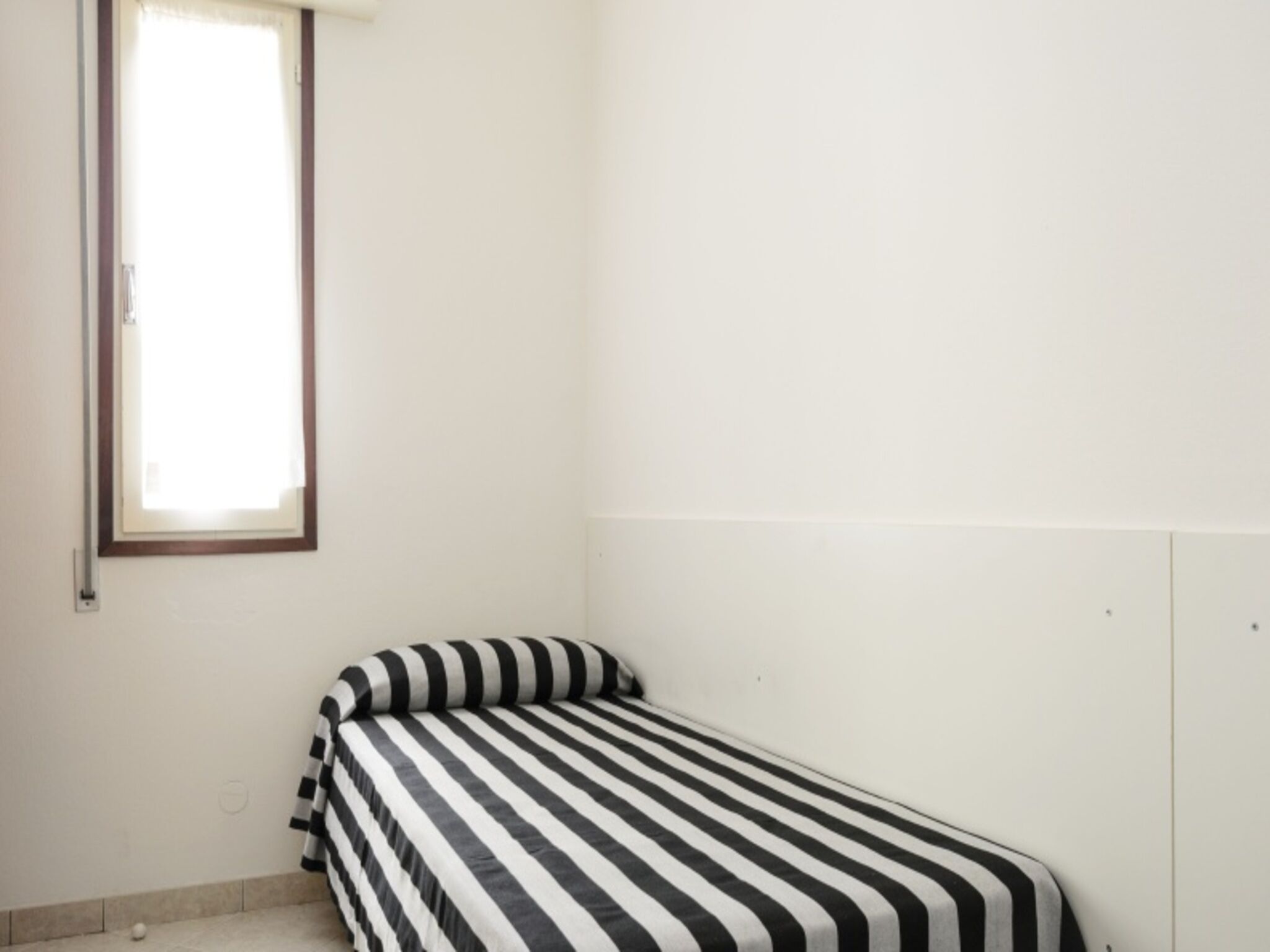 Ein kleines Zimmer mit einem Einzelbett, einem Fenster, schlichten Wänden und einem Bett mit gestreifter Bettdecke.