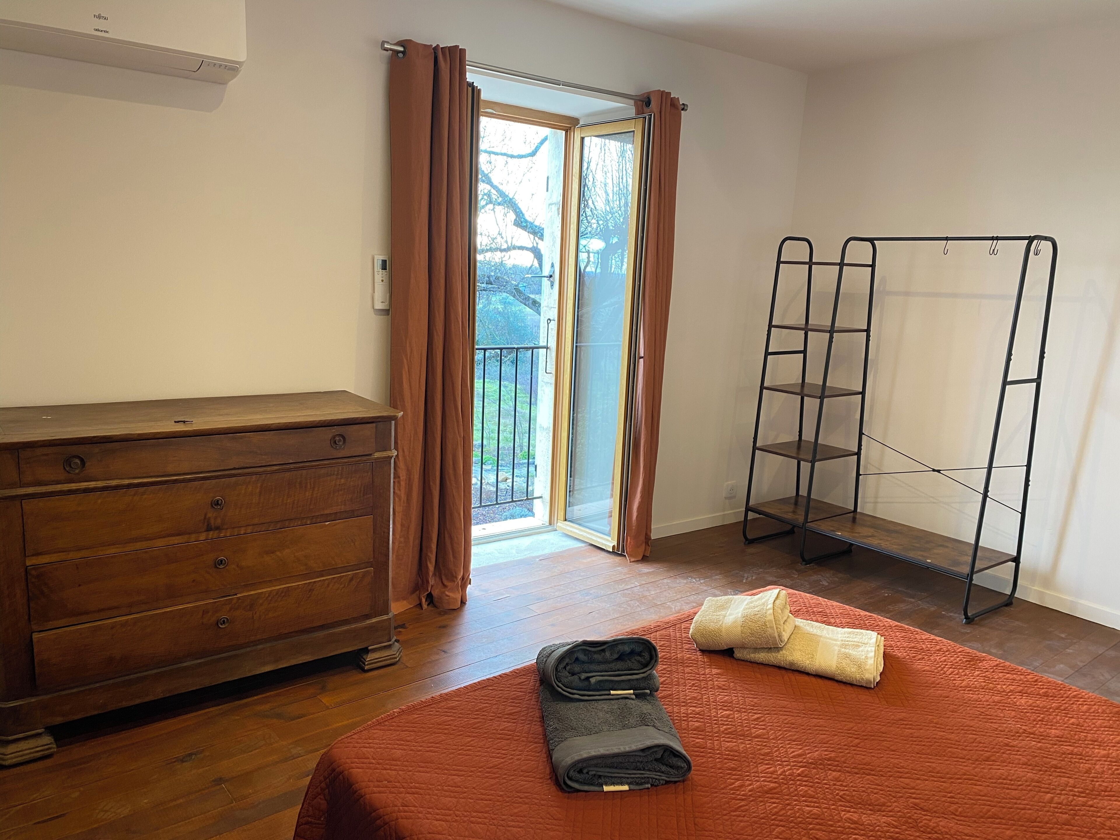 Un dormitorio con un aparador de madera, una cama con colcha roja, una estantería metálica y un ventanal corredero que da a un balcón.