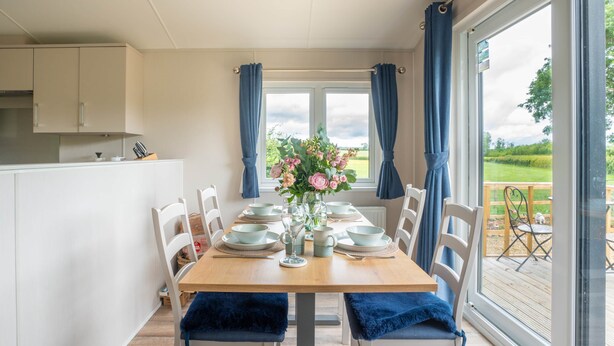 A dining area with a table set for six, chairs with blue cushions, a vase with flowers, and a view of the outdoors through large windows.