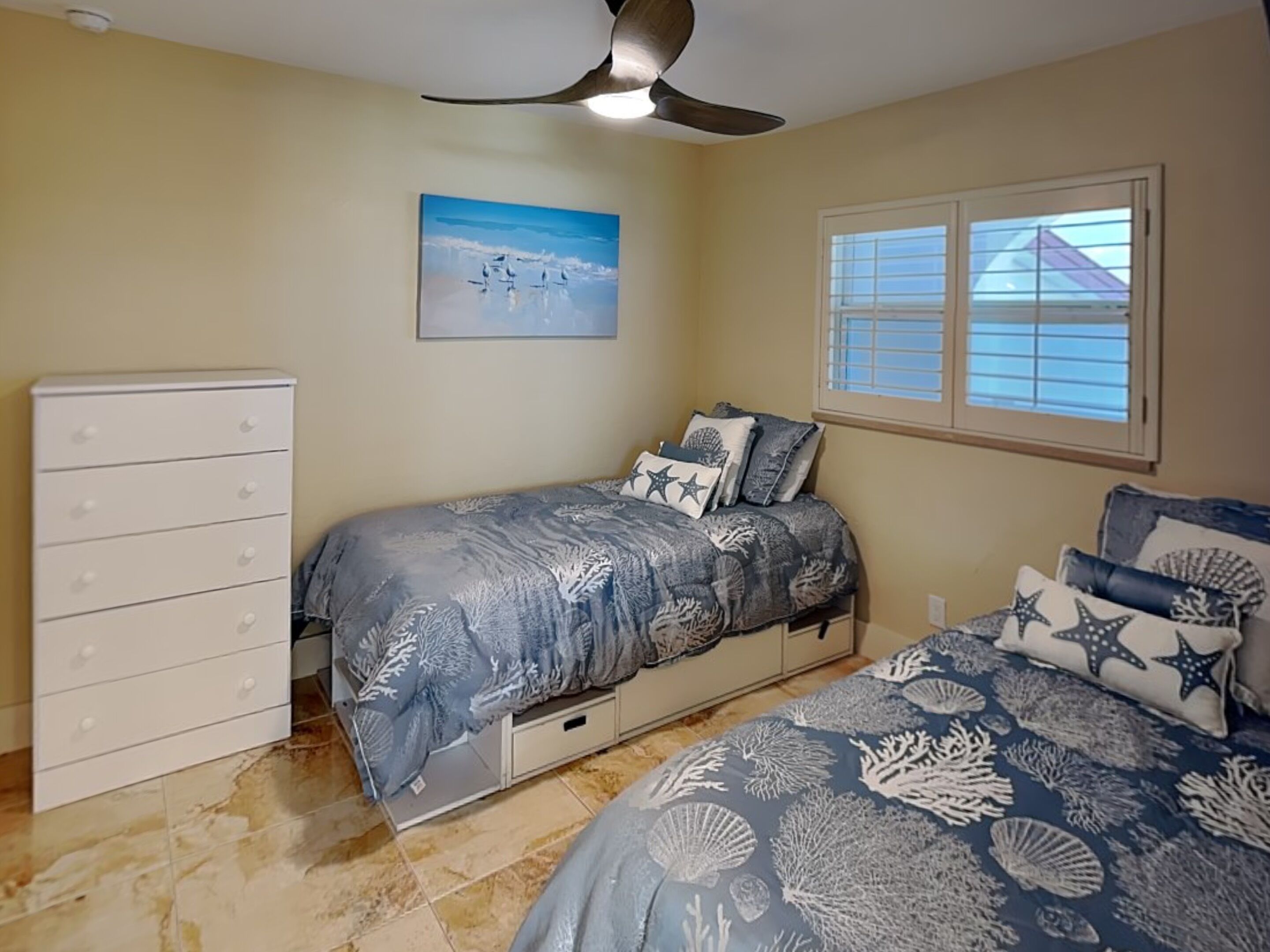 A bedroom with a bed, dresser, ceiling fan, painting, window, and tiled floor.