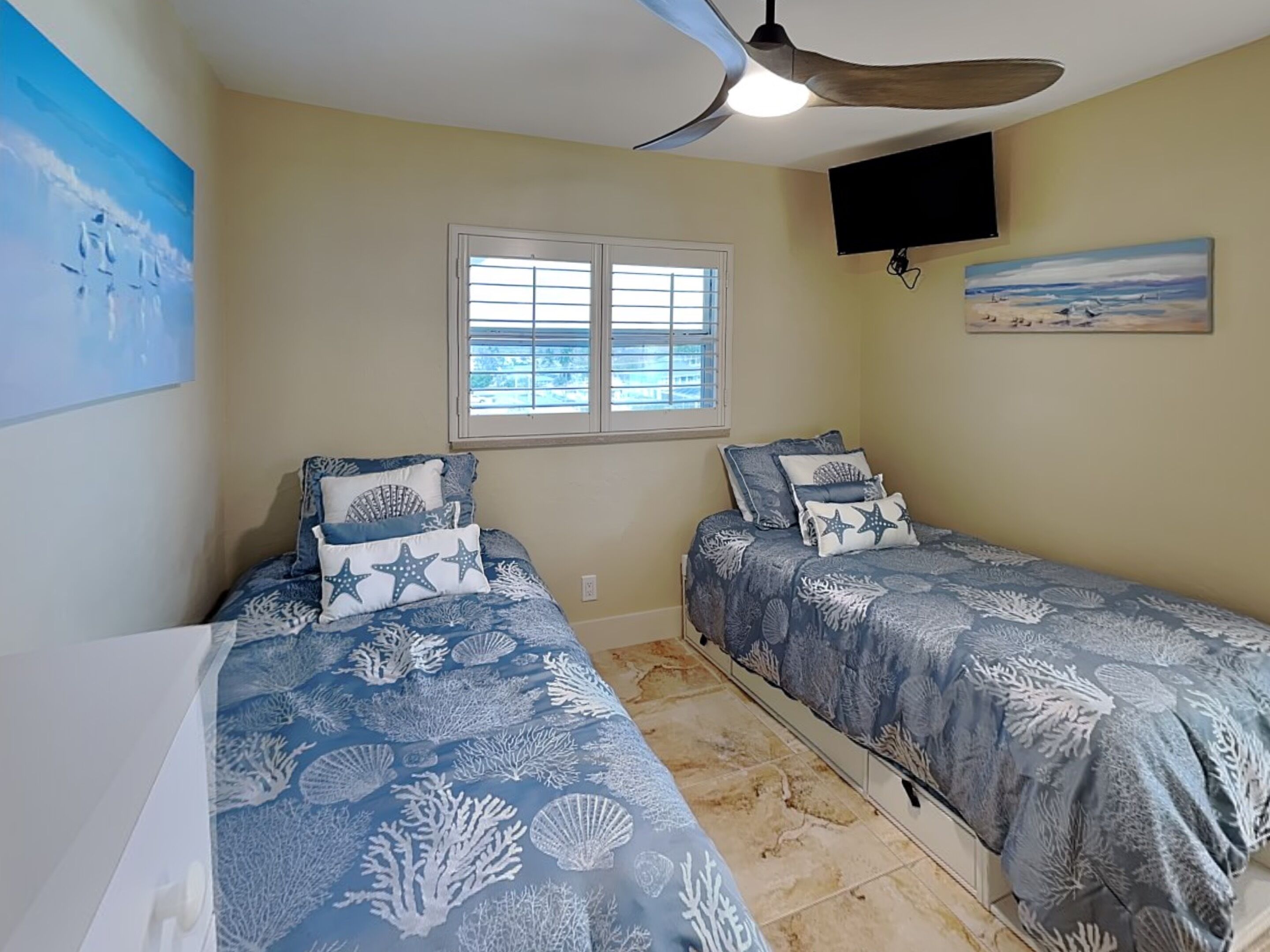 Une chambre avec deux lits, un ventilateur de plafond, une télévision, une fenêtre et des œuvres d’art accrochées aux murs.