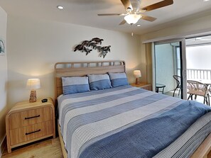Ein Schlafzimmer mit einem Bett, einem Nachttisch, einem Deckenventilator, Wandkunst mit Schildkröten und Blick auf einen Balkon mit Gartenmöbeln.