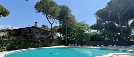 A swimming pool with a tiled deck, surrounded by trees and a building with a chimney.