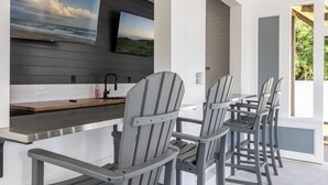 Et moderne udendørs køkken med barområde, grå Adirondack-stole og et tv på væggen, der viser et strandmotiv.