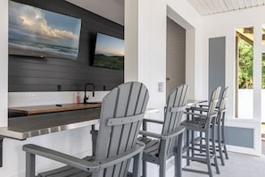 Eine moderne Außenküche mit Barbereich, grauen Adirondack-Stühlen und einem an der Wand montierten Fernseher, der eine Strandansicht zeigt.