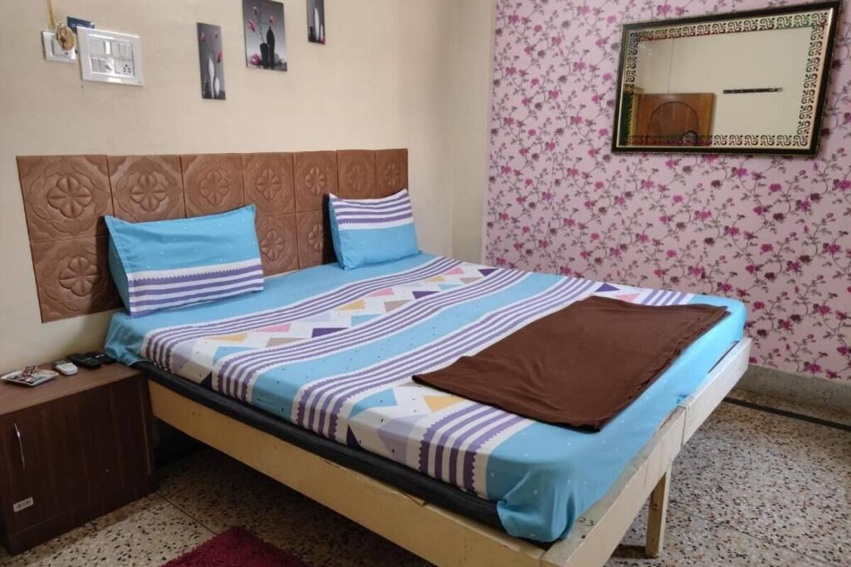 A single bed with a patterned bedspread, a wooden headboard, a nightstand, a red rug, and a wall with floral wallpaper and a mirror.