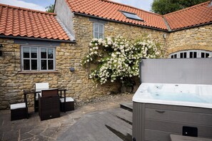 Een stenen gebouw met pannendak, een hottub, rieten meubels en bloeiende witte planten.