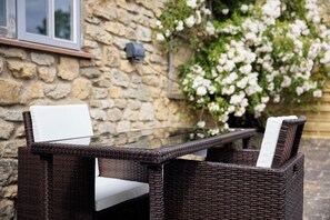 A wicker outdoor table with white cushions, set against a stone wall with a window and flowering plants. - Jasmine Cottage | Sands Farm (Wilton)