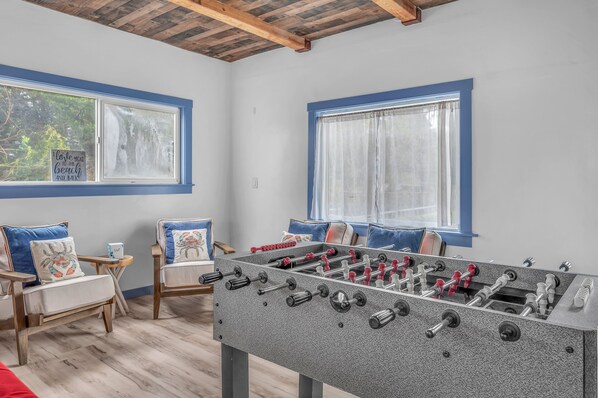 A room with a foosball table, two windows with blue frames, and a wooden ceiling. - Lincoln City Retreat: Private Hot Tub, Game Room, Easy Beach Access (Lincoln City)