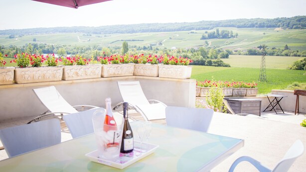 Eine Terrassengruppe im Freien mit Tisch, Stühlen, einer Weinflasche und Blick auf sanfte Hügel.