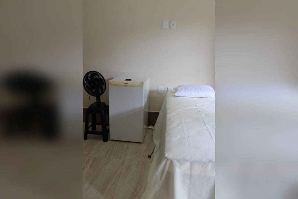 A small, simple room with a bed, a desk, and a fan.