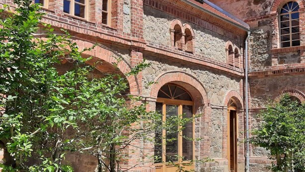 Un bâtiment historique doté de fenêtres en arc de cercle et d’une porte en bois, entouré d’arbres.