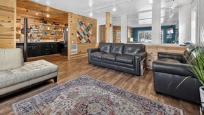 Ein modernes Wohnzimmer mit Sofa, Ledersessel, Teppich und Küchenbereich, der von Holzwänden sowie einem farbenfrohen Wandbild mit Rautenmuster geschmückt ist.