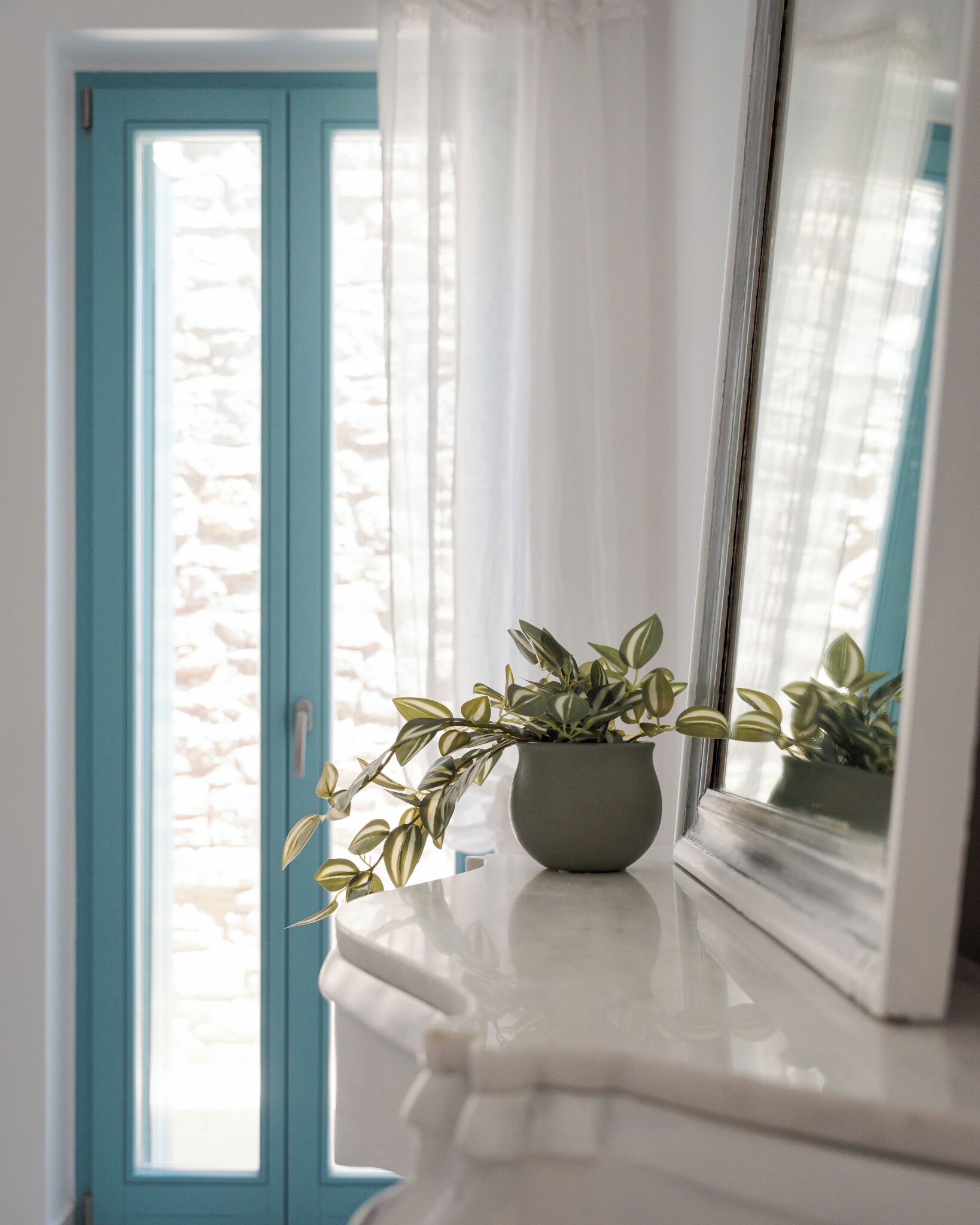 A potted plant on a white marble surface near a window with a blue door and a mirror reflecting the window.